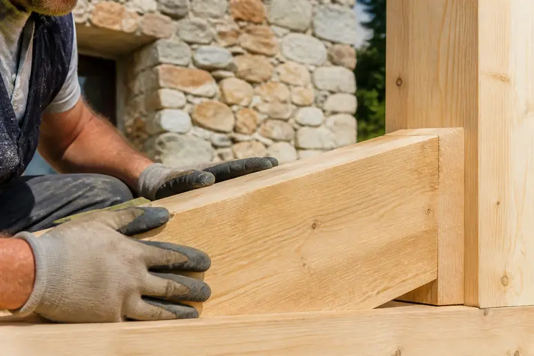 Mains d'ouvrier assemblant une maison bois et pierre, soulignant l'aspect écologique.
