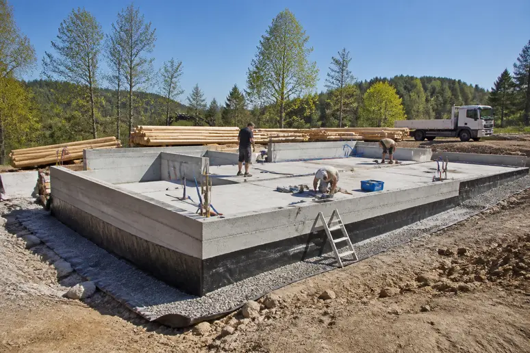 Chantier de construction d'une maison en rondins de bois avec ouvriers et fondations visibles.